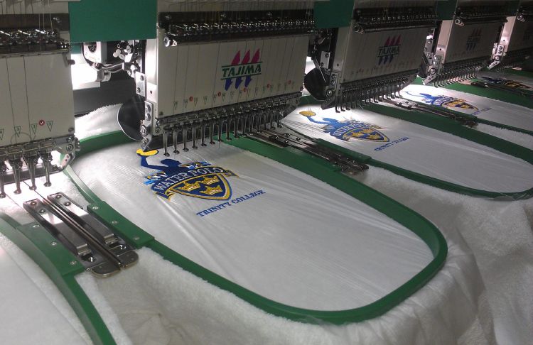 Multiple heads of a Tajima embroidery machine creating custom blue and yellow logos on white fabric.